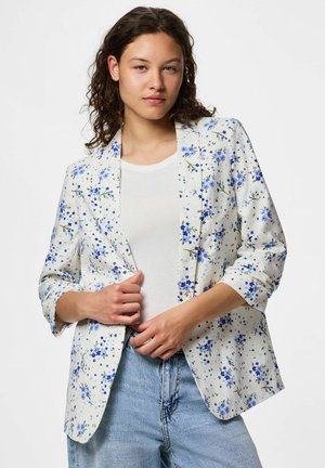 Tânără cu păr creț purtând un sacou alb cu imprimeu floral peste o cămașă albă și blugi bleu deschis, pozând cu mâinile lângă marginile sacoului.