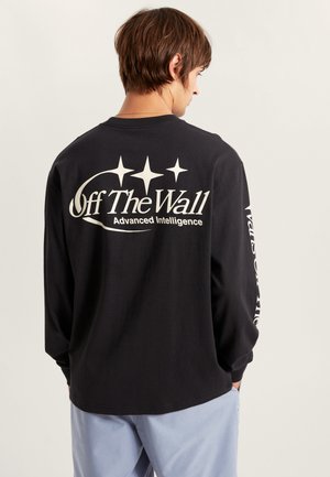 T-shirt noir à manches longues affichant du texte blanc et des graphismes d'étoiles au dos. Le tissu semble doux avec une coupe décontractée. Un pantalon bleu est visible.