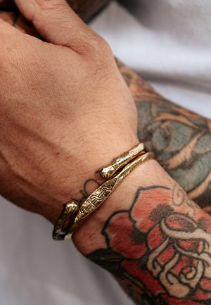 Close-up af en hånd og håndled med et tekstureret guldmanchetarmbånd og farverig blomster-tatovering på underarmen.