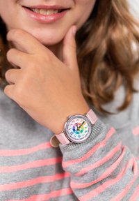 Montre d'enfant colorée avec un boîtier rond en métal argenté, un bracelet en cuir rose et un cadran numéroté arc-en-ciel avec des repères horaires clairs.