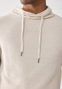 Hoodie beige clair en tissu tricoté texturé avec une capuche à cordon de serrage et des embouts métalliques argentés sur les cordons.