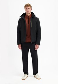 Zwarte gewatteerde jas met capuchon, ritssluiting aan de voorkant en zijzakken over een roestkleurige pullover. Zwarte broek en witte sneakers maken de look compleet.