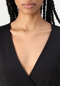 Svart v-ringad plagg med en jämn textur, som visar en djup halsringning och rena linjer, accentuerat av subjektets hudton.