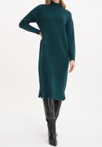 Lång teal färgad tröjklänning gjord av mjukt stickat material, med hög krage, långa ärmar, sidofickor och ribbade ärmslut och fåll.