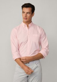 Camicia button-up di un delicato rosa chiaro con taschino sul petto, maniche arrotolate e una texture in cotone morbido, abbinata a pantaloni grigi chiari.