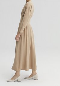 Beige midi šaty s dlouhými rukávy a kulatým výstřihem; mají splývavou látku a skladby; jsou doplněny světle béžovými lodičkami na blokovém podpatku.