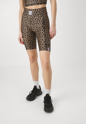 Person iført leopardmønstrede high-waist biker shorts med sorte sneakers og hvide sokker, mod en ensfarvet lys baggrund.