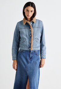 Jeansjacka med en beige, fluffig krage, som har en knäppning fram, två framfickor och en kort design, ihop med en blå jeanskjol.