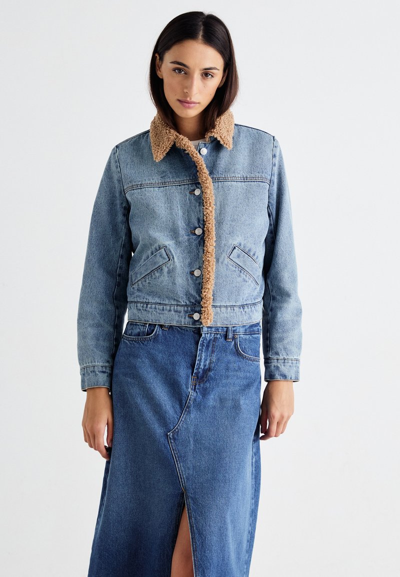 Jeansjacka med en beige, fluffig krage, som har en knäppning fram, två framfickor och en kort design, ihop med en blå jeanskjol.