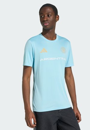 Ung mand iført en lyseblå Argentinsk fodboldtrøje med Adidas-logo og sorte jeans, ser sidelæns mod en ensfarvet baggrund.
