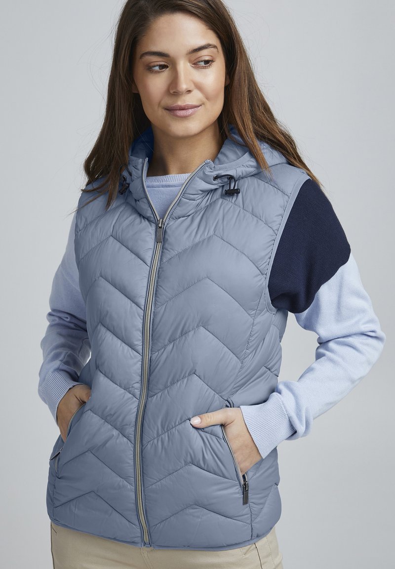 Vest matlasat de culoare albastru deschis, cu glugă, dotat cu fermoar frontal, buzunare laterale și accente tricotate de culoare navy pe umeri și mâneci.