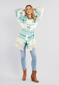 Beige kimono med turkosa geometriska mönster, fransar i nederkanten och bälte; kombinerad med ljusblå jeans och bruna ankelskor.