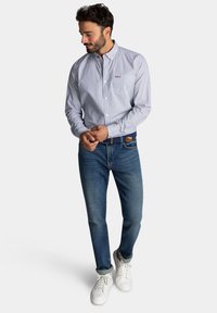 Blauw en wit gestreepte button-up blouse, slim-fit blauwe jeans en witte sneakers. Opvallende details zijn een gebrandmerkt logo op de borst en opgerolde mouwen.