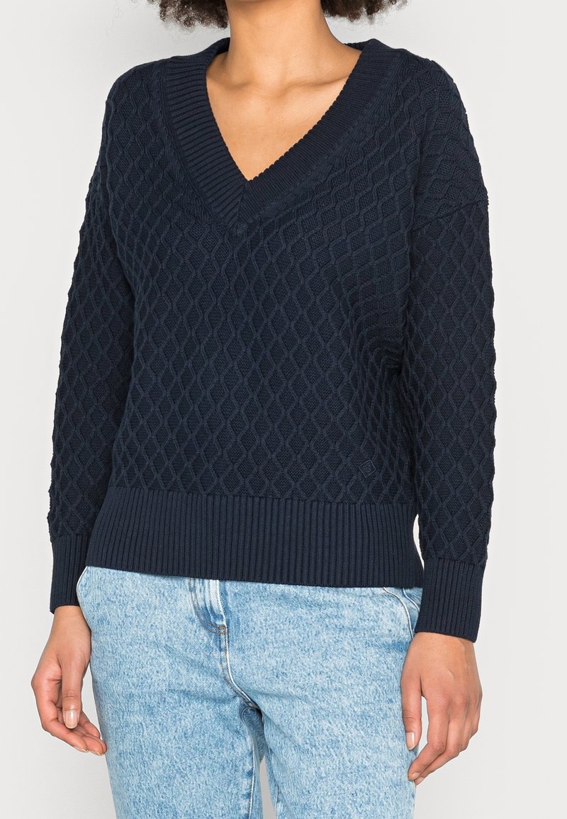 Femme portant un pull à col en V texturé bleu marine et un jean bleu clair, debout avec les bras détendus le long du corps.
