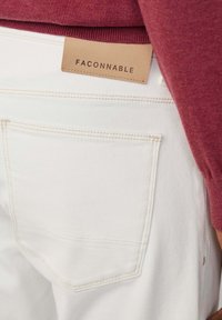 Pantalon en denim blanc avec une poche arrière visible et une étiquette en cuir marron clair portant le nom de la marque "FACONNABLE" cousue sur la taille.