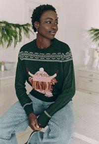Dunkelgrüner Pullover mit einem Design einer Highland-Rinderkuh, die einen rosa Hut trägt, verziert mit glänzenden Akzenten und Fransen. Kombiniert mit hellblauen Jeans.