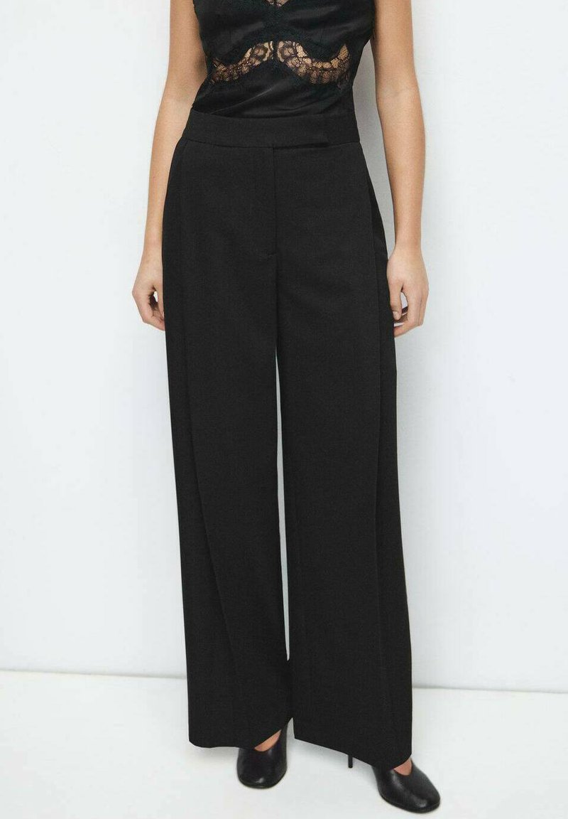 Pantaloni negri cu picior larg și talie înaltă, confecționați dintr-un material neted și cu cusături minimale, asortați cu un top negru din dantelă și pantofi cu toc înalt.