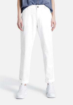 Vrouw met opgerolde witte broek, witte sneakers en een blauwe blouse die is ingestopt, staande tegen een effen lichte achtergrond.