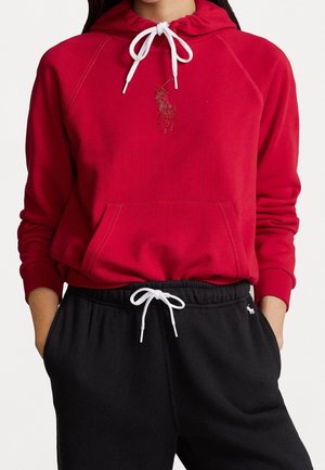Kobieta ubrana w czerwoną bluzę z kapturem z białymi sznurkami oraz czarne spodnie dresowe z białym sznurkiem, oba elementy z małymi haftowanymi logo gracza polo.