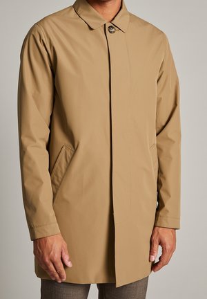 Homme portant un manteau beige clair à un seul bouton avec des poches latérales et des manches longues, associé à un pantalon marron.