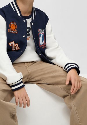 Kind trägt marineblaue und weiße Collegejacke mit Basketball-Patches, weißes Hemd und beige Hose, sitzt mit den Händen auf den Beinen.