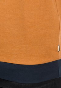 Pull orange avec un détail d'ourlet bleu marine porté sur un pantalon noir, montrant une petite étiquette en tissu sur la couture latérale.