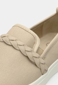 Chaussure beige à enfiler en tissu texturé avec un accent tressé le long du dessus et une semelle blanche. Design à bout arrondi.