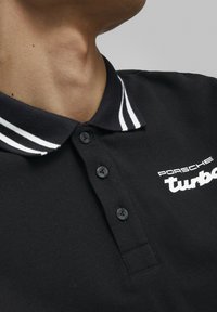 Svart polo skjorte med strukturert stoff, tre svarte knapper, hvite krage-striper og brodert "Porsche turbo"-logo i hvitt.