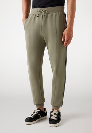 Joggers verde olivo de corte tapered, con cintura ajustable, rayas laterales y puños de canalé en los tobillos. Combinados con zapatillas negras.
