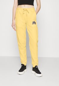 Gelbe Sweatpants mit einem Kordelzug an der Taille, versehen mit einem blauen gestickten Logo am linken Oberschenkel. Das Material wirkt weich und hat einen lockeren Schnitt.