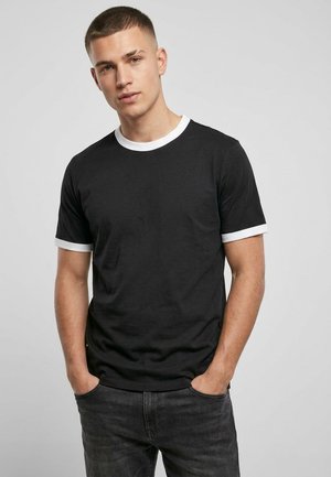 Junger Mann trägt schwarzes T-Shirt mit weißem Kragen und Ärmelsäumen, steht mit Händen in den Taschen vor einfarbigem grauem Hintergrund.