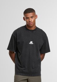 Mladý muž s krátkými vlasy, oblečený v volném černém tričku s malým bílým logem a khaki kalhotami, stojící s rukama za zády.