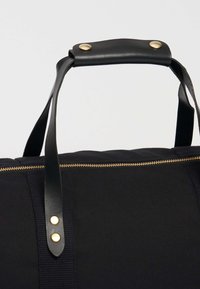 Černá látková taška s černou koženou rukojetí, vybavená mosaznými komponenty a zlatým zipem na vrchu. Texturovaná látka a hladké kožené detaily.