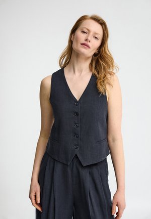 Vrouw met een lichte huid en rood haar draagt een mouwloos donker button-vest en bijpassende hoge taillebroek, staand tegen een effen achtergrond.