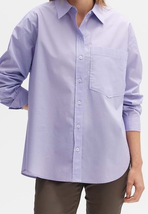Femme portant une chemise ample mauve clair à boutons avec une poche poitrine et des manches retroussées, associée à un pantalon foncé.