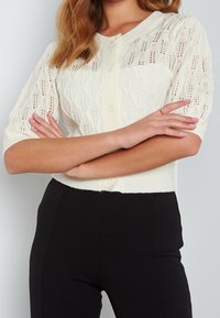 Pletený krémový cardigan s krátkými nařasenými rameny, se vzorem v cable knit, kulatým výstřihem a zapínáním na knoflíky vpředu.