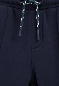 Tamsiai mėlynos treninginės kelnės su tekstūruota medžiaga, turinčios raištelį su žalia, balta ir mėlyna juostelėmis ir šonines kišenes.