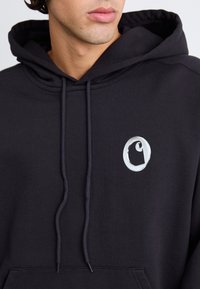Sudadera negra con capucha, hecha de algodón, con capucha ajustable y un logo blanco circular en el pecho. Textura suave y corte relajado.