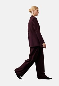 Bordeaux op maat gemaakt pak met een getailleerd jasje en wijde broekspijpen. Glad stof met een matte afwerking. Zwarte schoenen en gouden oorbellen als accenten.