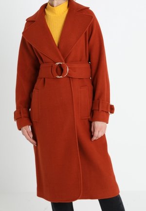 Rust orange belted wool coat with wide lapels and cuff straps worn over a yellow turtleneck and black pants.