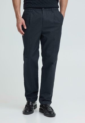 Mann som har på seg svarte, smale bukser og svarte skinnloafers, stående med hendene i lommene foran en ensfarget bakgrunn.