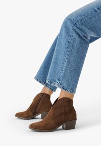 Brązowe zamszowe botki na blokowym obcasie noszone z niebieskimi dżinsami na białym tle.