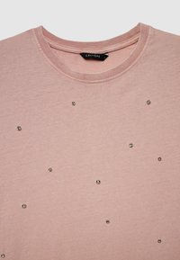 Hellrosa kurzärmeliges T-Shirt aus weicher Baumwolle, mit verstreuten metallischen Nieten auf der Vorderseite und einem runden Ausschnitt.