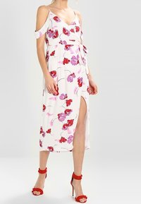 Robe midi florale blanche ornée de motifs de fleurs rouges et violettes, avec des épaules dénudées et une fente sur le côté. Ceinturée à la taille, accompagnée de sandales à talons rouges.