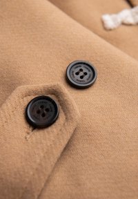 Hnědá vlněná látka s hladkou texturou se vyznačuje dvěma černými knoflíky a designem šikmého klopového kapsy. Detaily knoflíků zahrnují čtyři dírky.