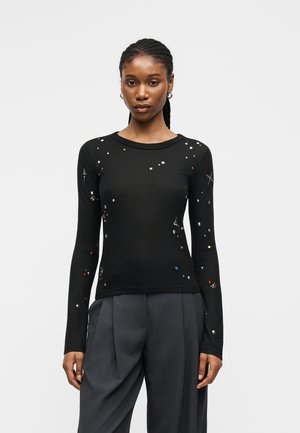 Noor naine palmikutega, kes kannab musta pika varrukaga toppi väikeste hajutatud kaunistustega ja lahtisi halli volditud pükse.