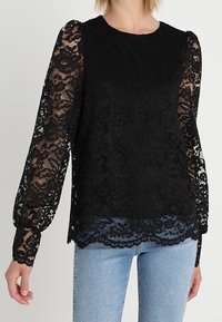 Top in pizzo nero con maniche lunghe e a sbuffo, con orlo sagomato e motivi floreali. Abbinato a jeans in denim azzurro chiaro.