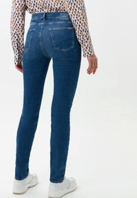 Džíny v úzkém střihu z modrého denimu s vysokým pasem, zadními kapsami, jemným zesvětlením a jemnými detaily prošití, spárované s bílými sportovními botami.