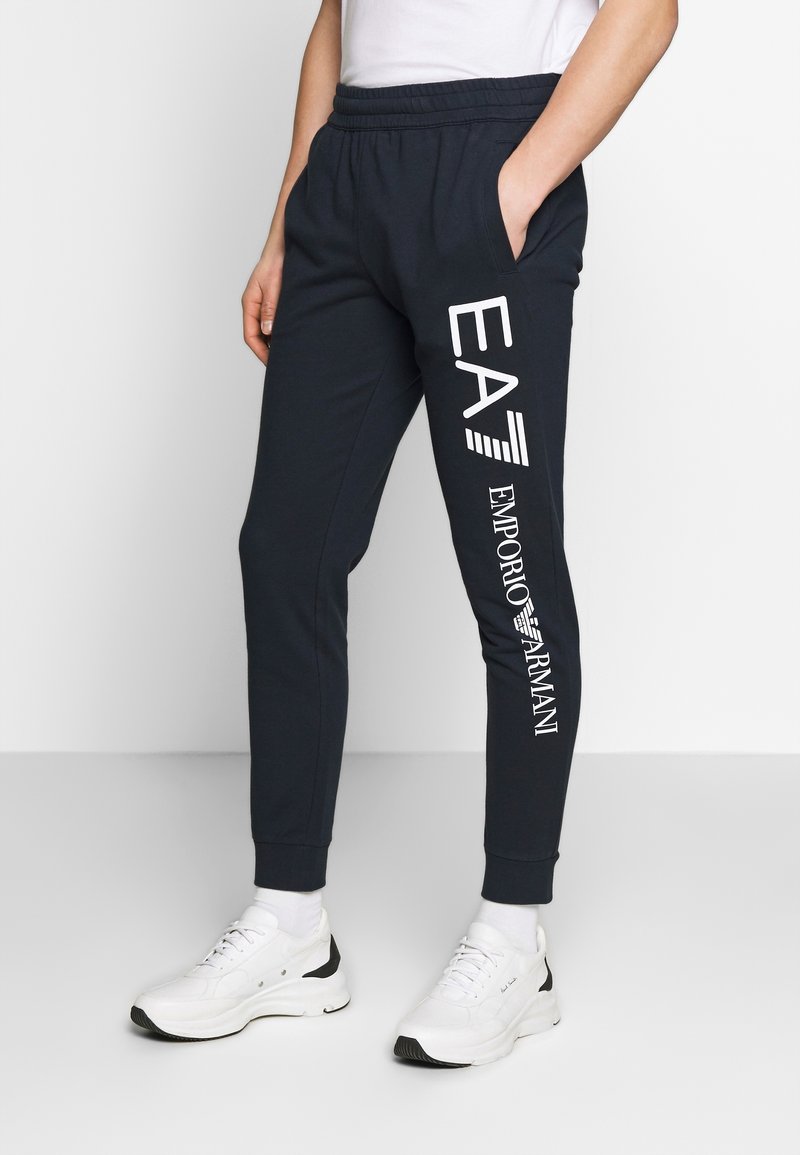 Námořnické tepláky s elastickým pasem, bočními kapsami a bílým potiskem loga "EA7 EMPORIO ARMANI" na levé noze. Bavlněná směs tkaniny.