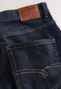 Tasca posteriore di jeans in denim blu scuro con cuciture gialle e un'etichetta di pelle marrone con la scritta "Next Signature Denim Goods."